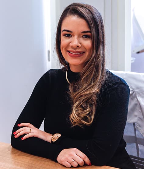 A imagem mostra uma mulher sentada a uma mesa, com os braços cruzados sobre a mesa. Ela está vestindo um suéter preto de manga longa e usa um relógio de ouro no pulso esquerdo. Seus cabelos são longos e castanhos, com mechas mais claras, e estão soltos sobre os ombros. Ela tem as unhas pintadas de vermelho. O fundo da imagem é um ambiente interno, possivelmente um consultório em Salvador, BA, com uma janela visível à esquerda e uma cadeira coberta com um pano branco à direita. A iluminação é suave e natural, provavelmente proveniente da janela. A mulher parece estar em uma postura relaxada e confiante.